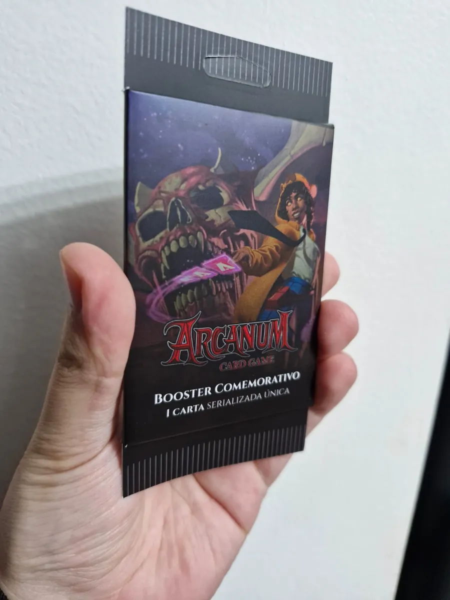 Mão segurando o Booster Comemorativo do Arcanum, com a arte da Nanda e uma caveira gigante, e o texto 1 carta serializada única
