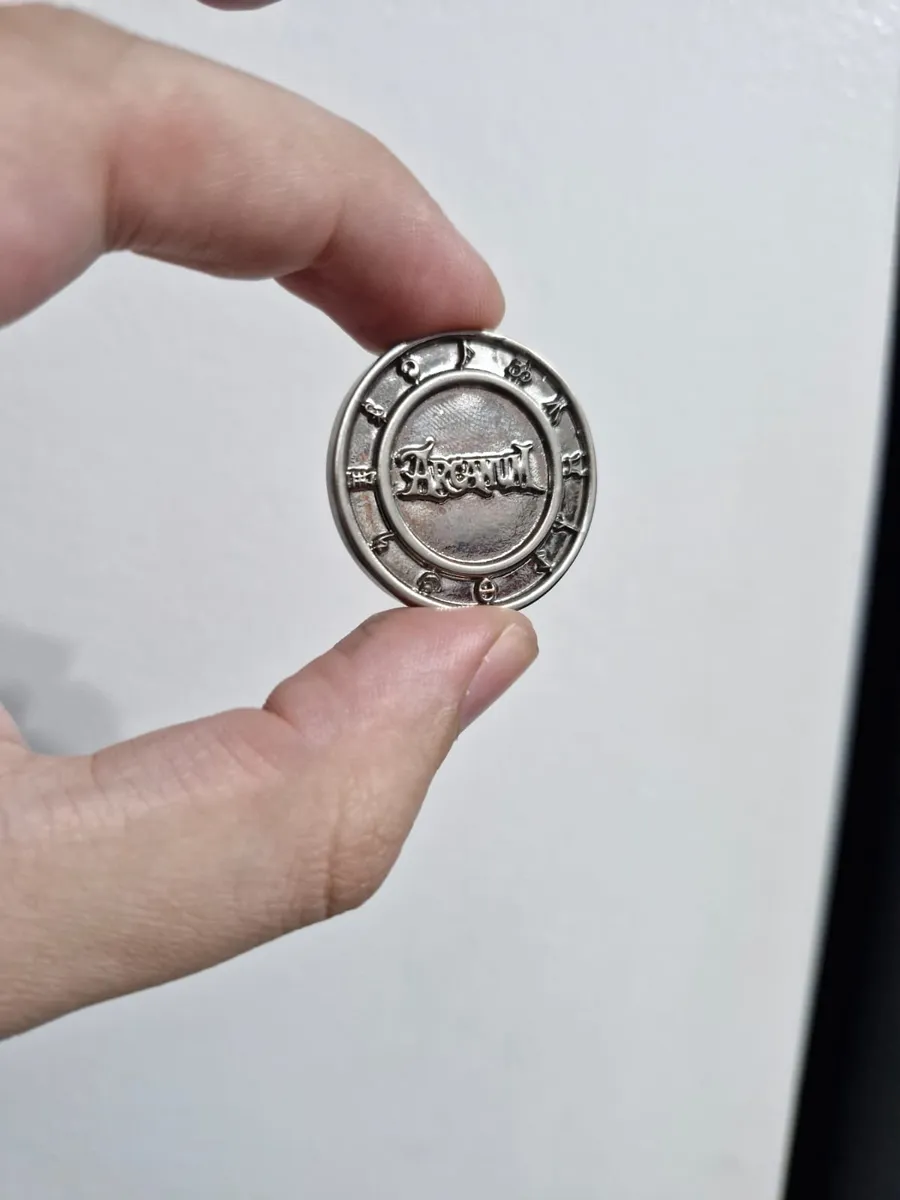Verso da moeda de metal com o logo do Arcanum em relevo