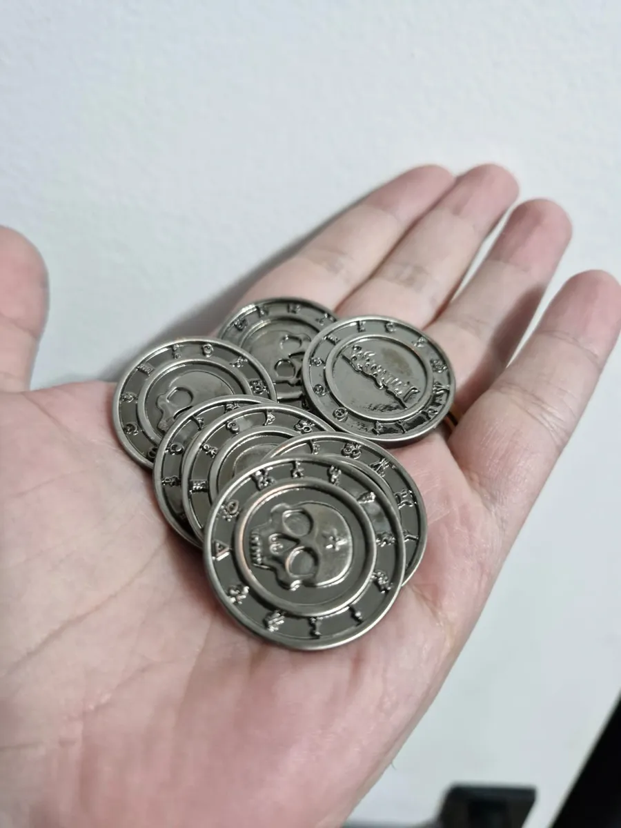 Várias moedas de metal prateadas do Arcanum na palma da mão, com caveiras e o logo em relevo