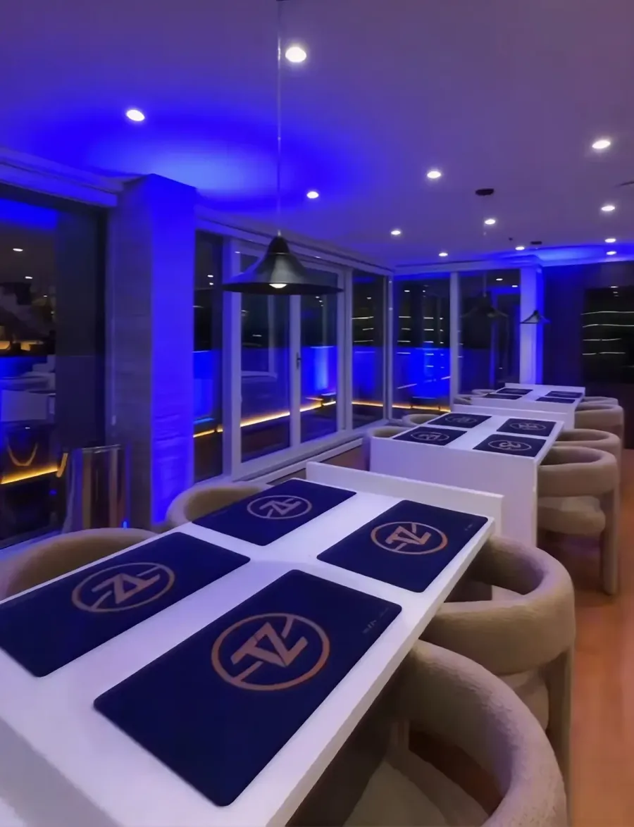 Salão da Tuzzy Cards Lounge com mesas brancas em fila, playmats azuis com o logo da Tuzzy e luz azul no ambiente