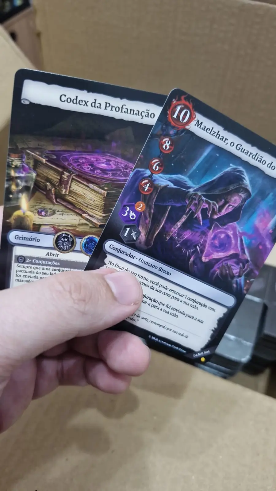 Mão segurando as cartas impressas Codex da Profanação e Maelzhar, o Guardião do Vazio