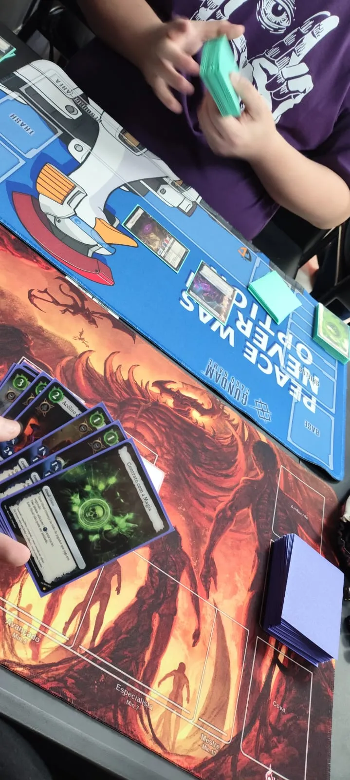 Vista de cima de uma partida, com uma mão de cartas do Arcanum sobre um playmat de arte flamejante