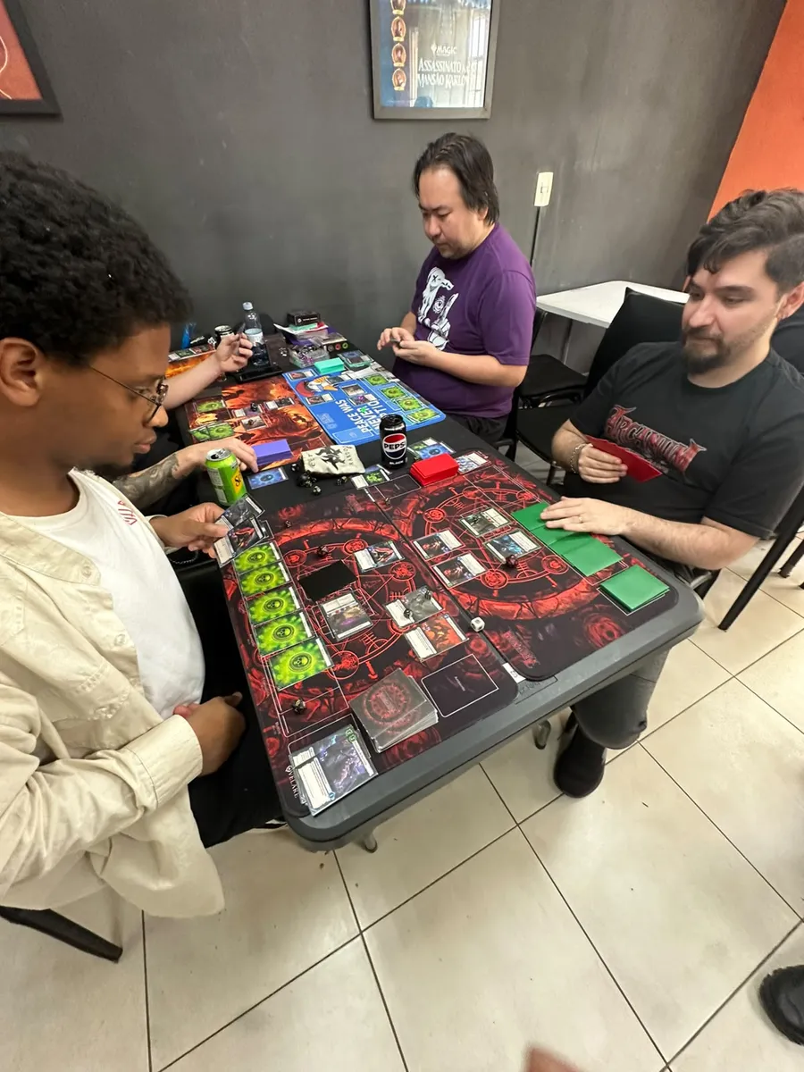 Jogadores concentrados em duas mesas da Guilda Geek Games, com playmats do Arcanum, cartas em jogo e dados sobre a mesa