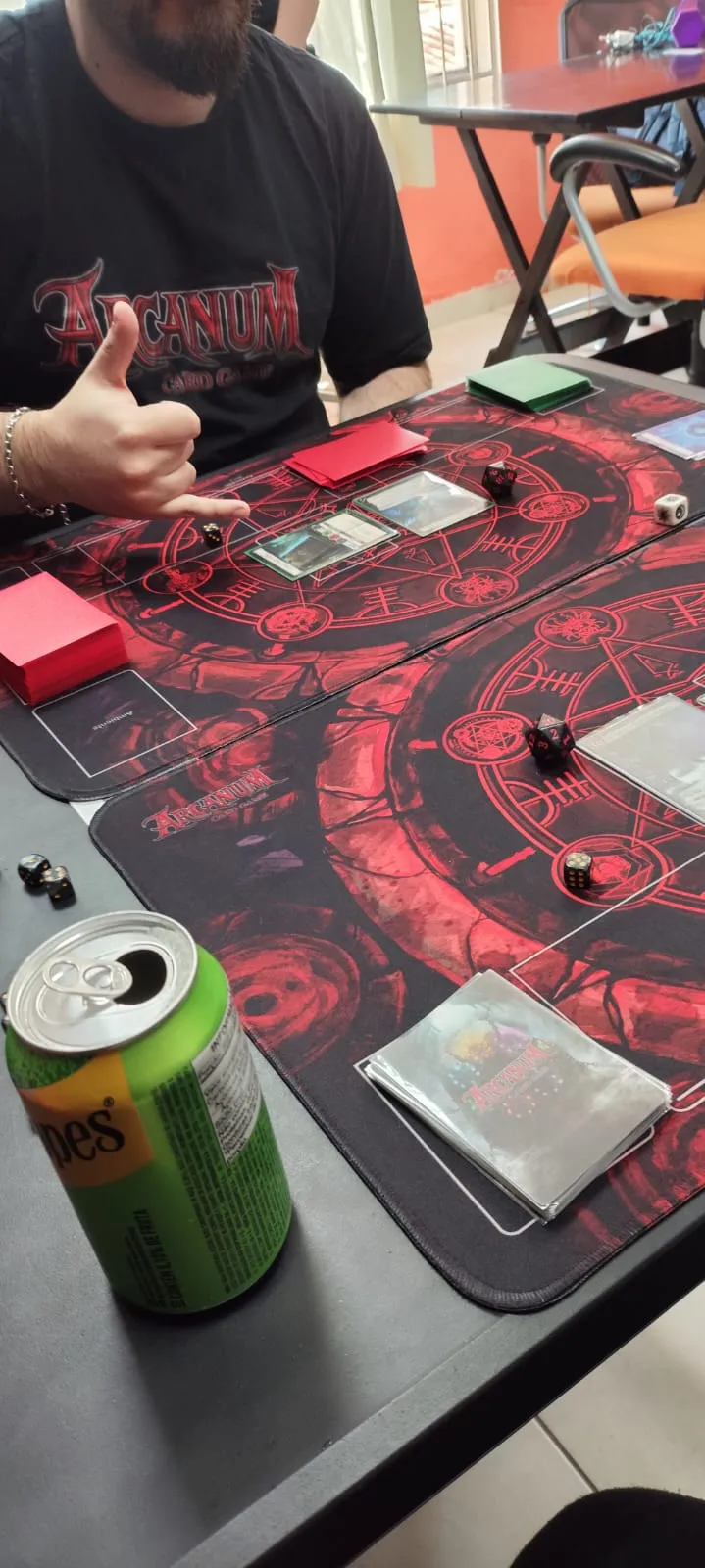 Jogador com camiseta do Arcanum fazendo sinal de positivo durante a partida, com cartas e dados sobre os playmats