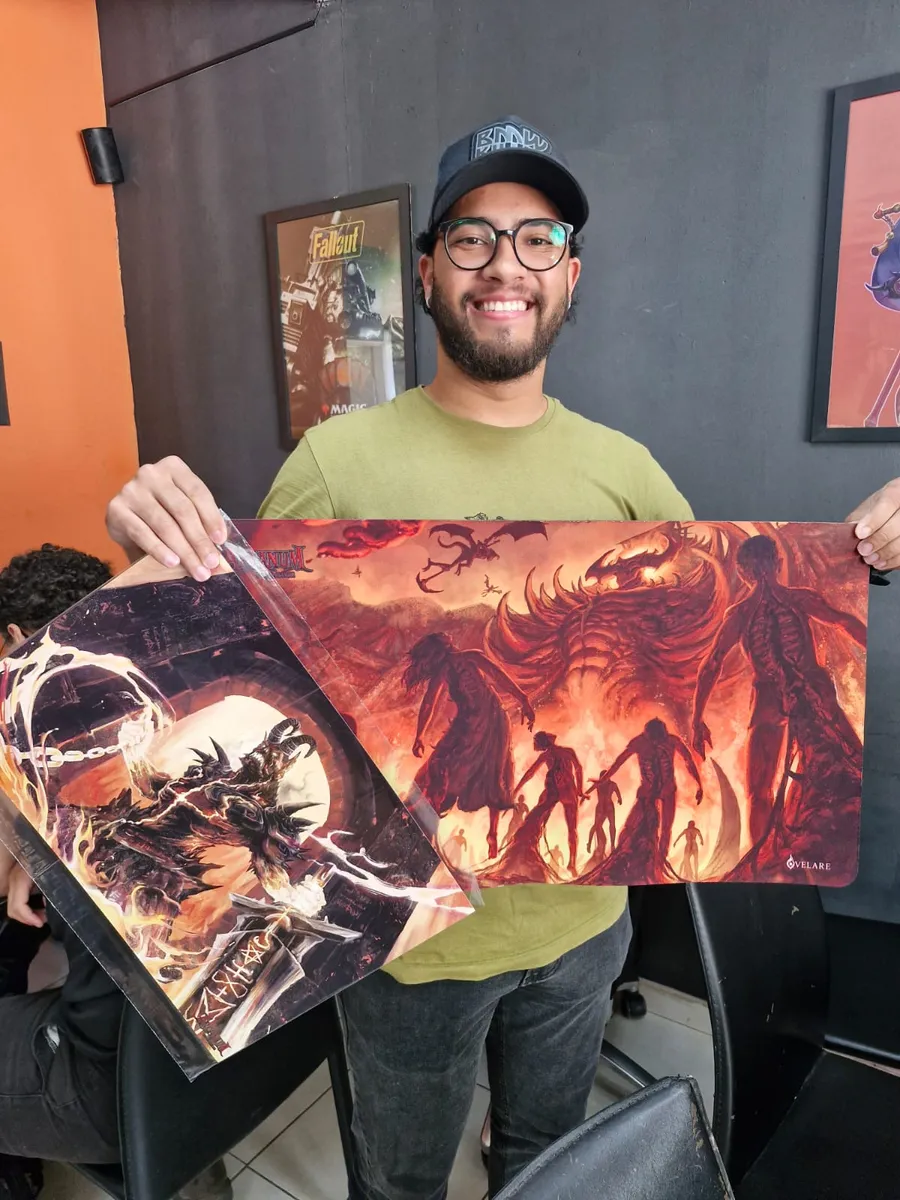 Jogador sorridente segurando dois playmats do Arcanum, um com arte de demônio acorrentado e outro com figuras caminhando em meio ao fogo
