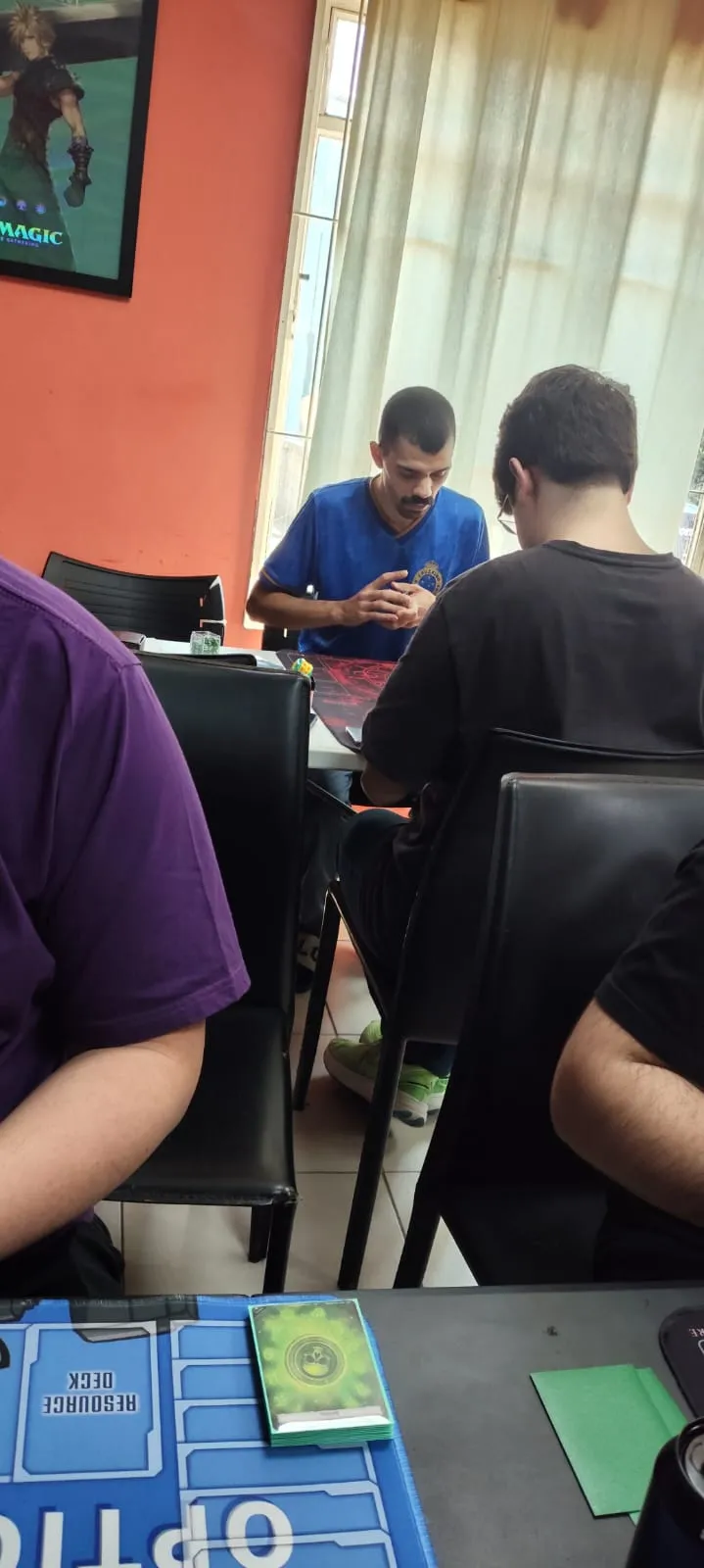 Dois jogadores se enfrentando numa mesa ao fundo da loja, perto da janela